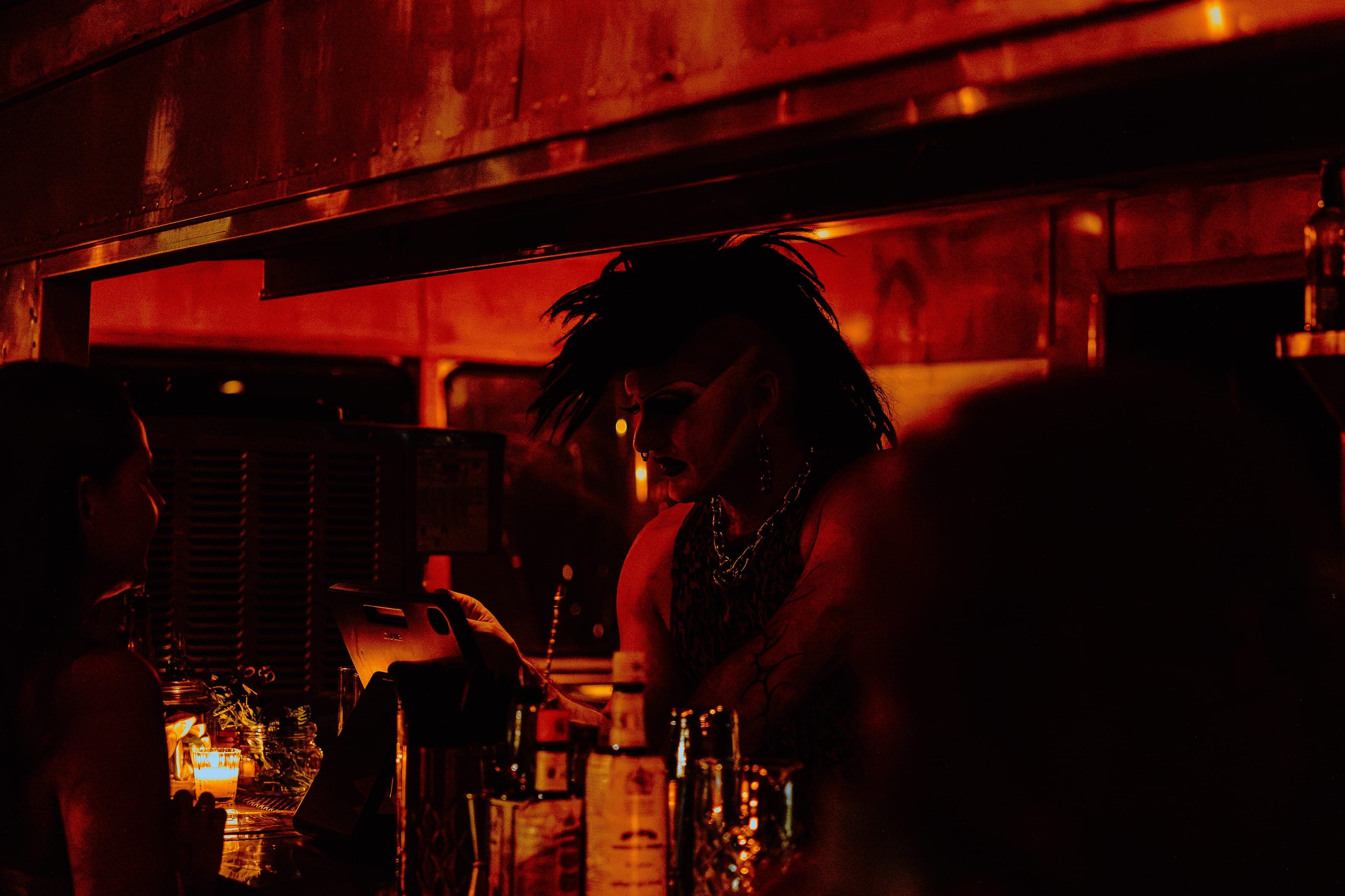 A bartender with a feathered black mohawk and chain necklace working the bar, bathed in red light
