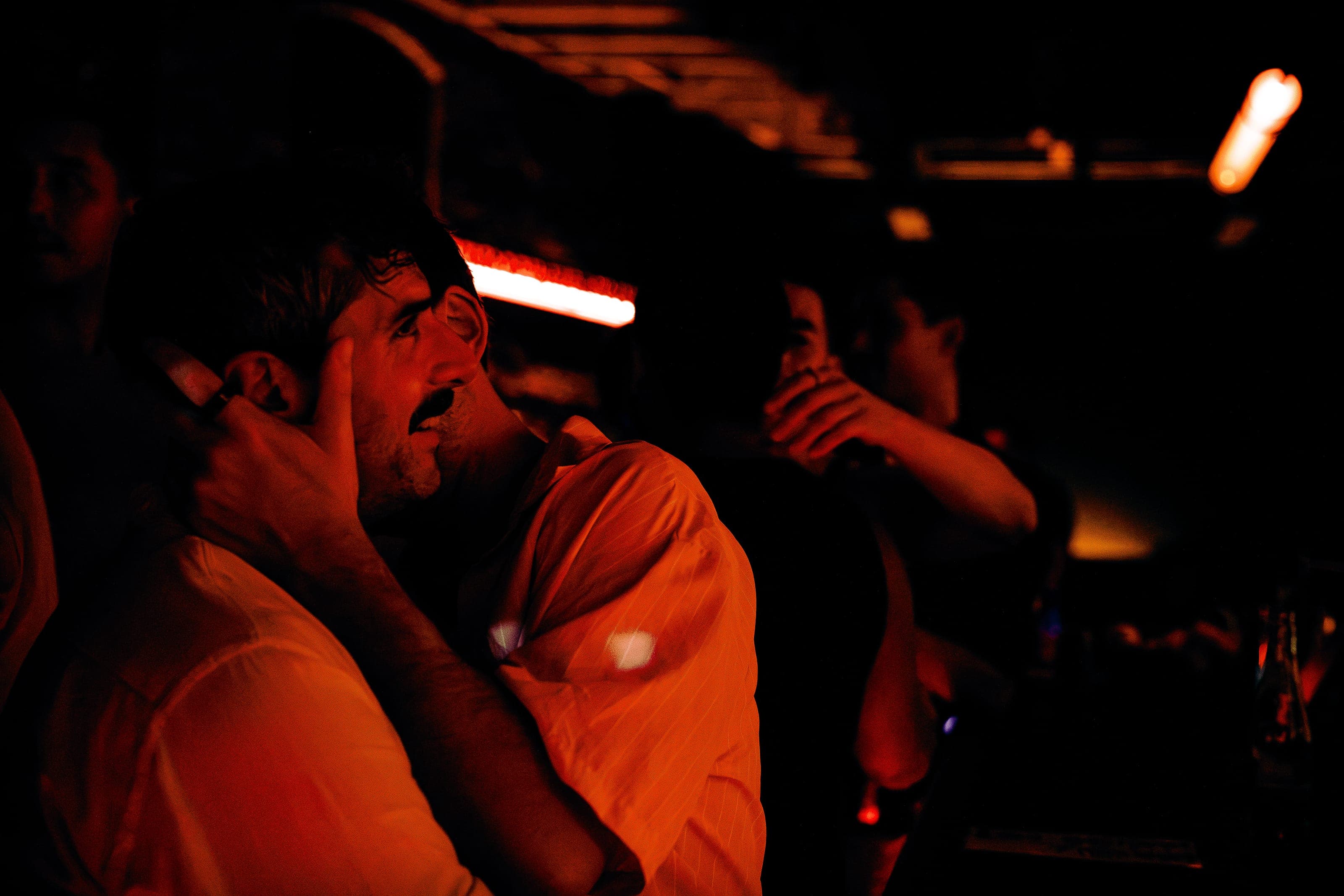 Two patrons close together at the bar, one with a hand to his face, in warm red light
