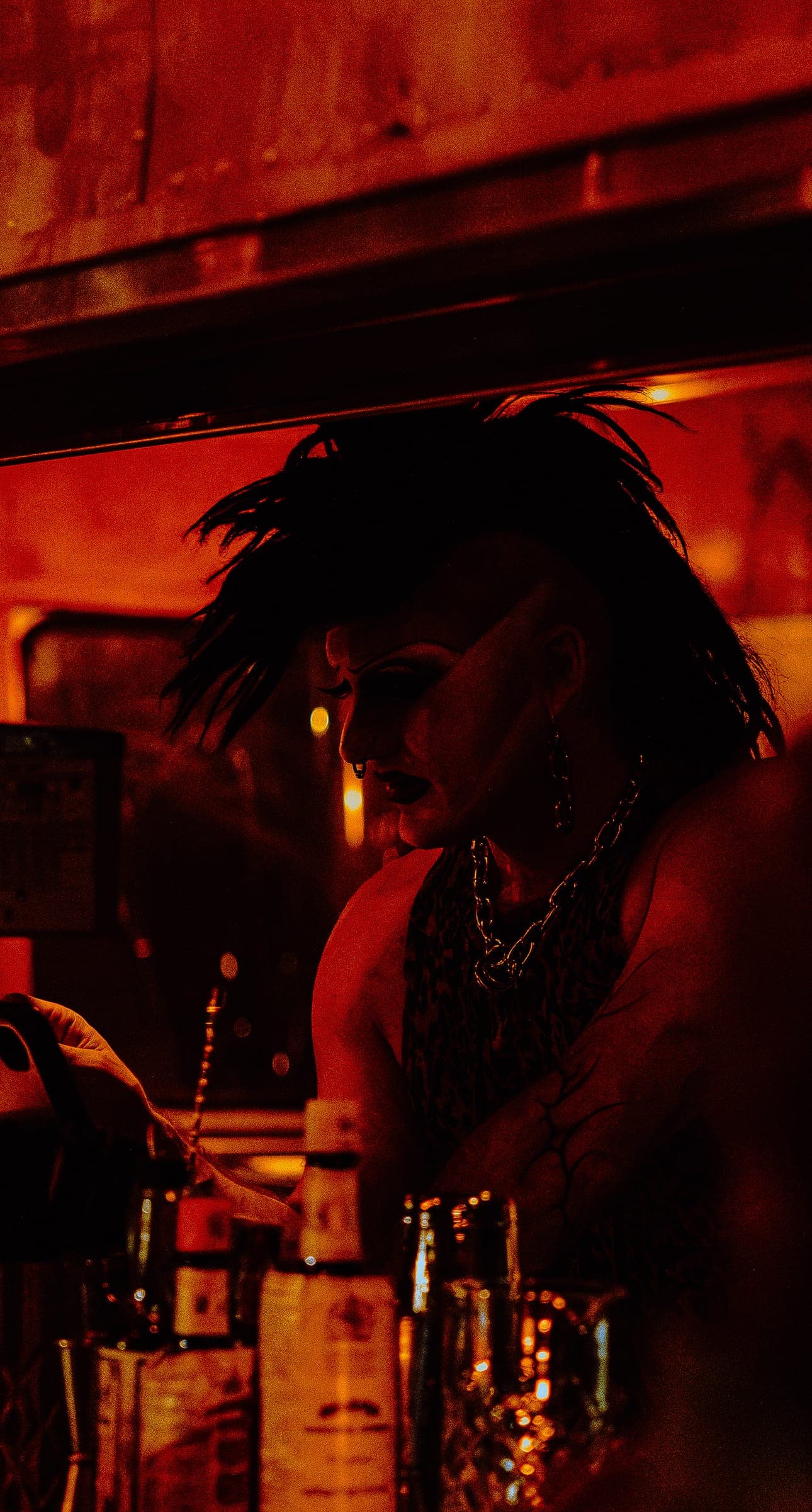 A bartender with a feathered black mohawk and chain necklace, bathed in red light