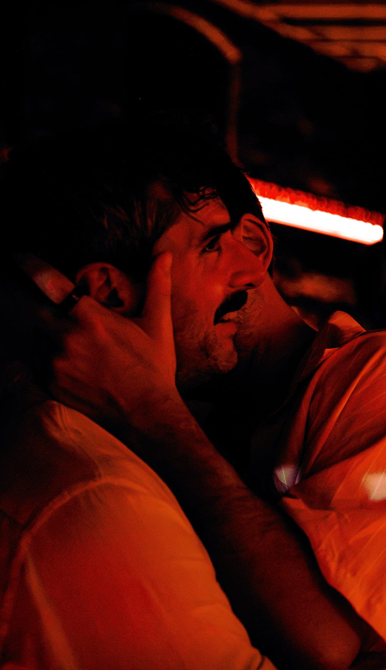 A patron with a hand to his face, close to someone at the bar, in red light